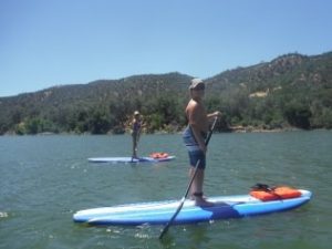 Two people paddleboarding on a calm lake surrounded by trees and hills. Sunny day.