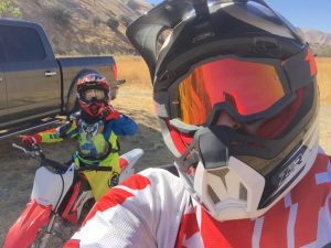 Father and son dirt biking, both wearing helmets and gear. Red Honda dirt bike.