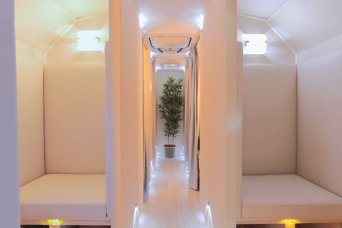 Minimalist nap pod interior with white seating, soft lighting, and a plant at the end of the hallway.