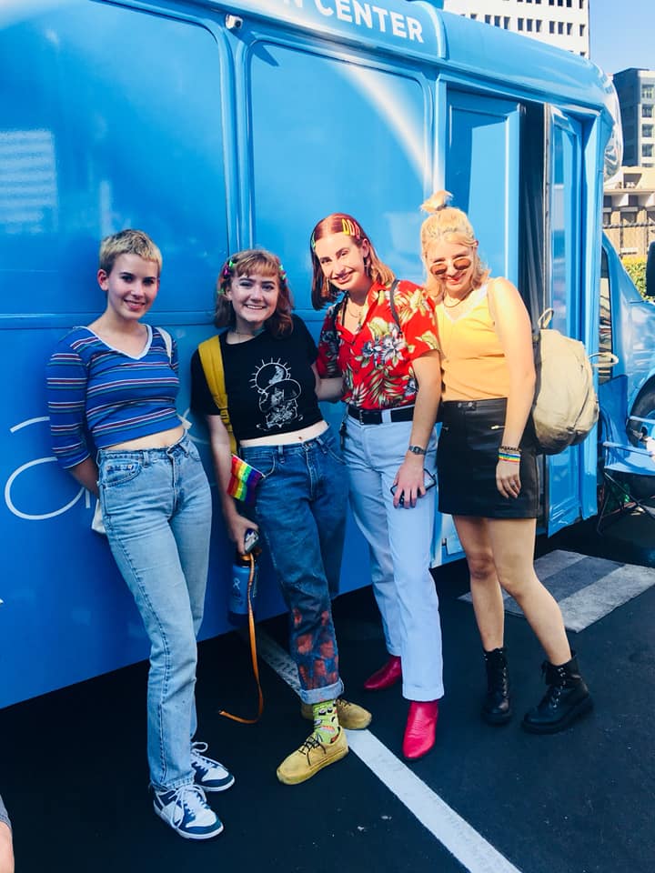 Four stylish friends pose by a blue van, showcasing diverse fashion and friendship.