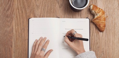Open planner, coffee, and croissant on a wooden desk for morning routine planning.