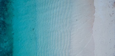 Aerial view of clear turquoise water meeting a white sand beach with subtle wave patterns.