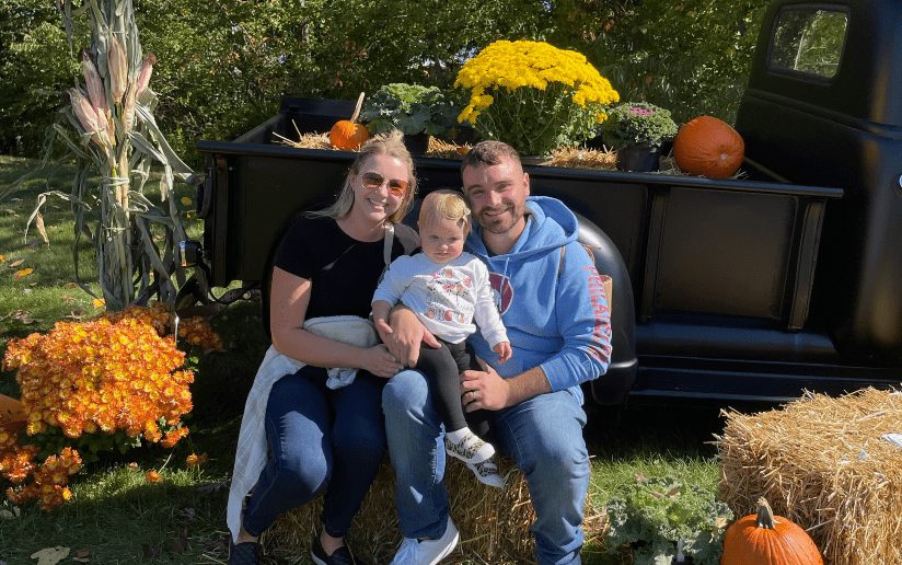 Andrew Albert with family at a fall festival, sitting on hay bales near a vintage truck with pumpkins and mums.