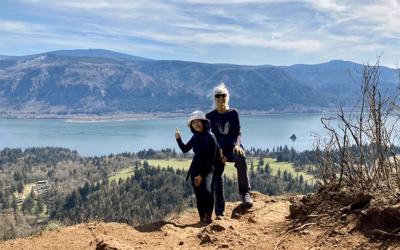 Two women pose on a scenic overlook with a river and mountains in the background.