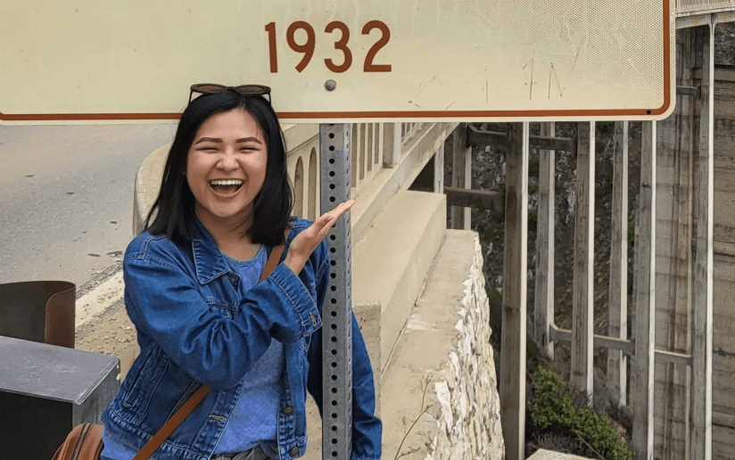 Jackie Tran stands by a sign reading "1932" at Bixby Bridge, smiling and pointing at the historic landmark.