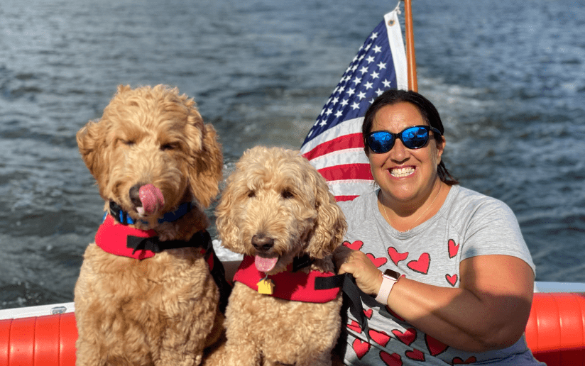 Michelle Harden with two Goldendoodle dogs wearing life jackets on a boat with an American flag.