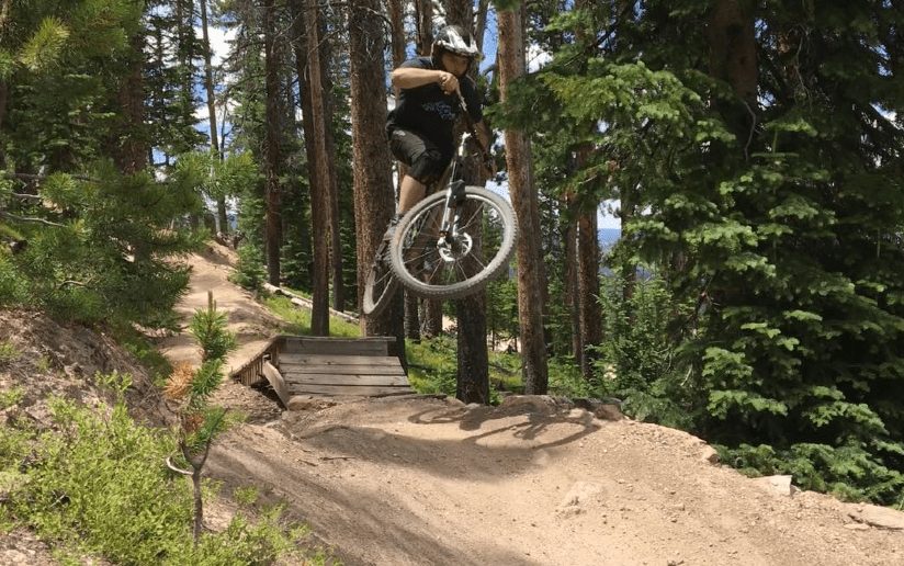 Mountain biker catching air off a wooden jump in a forest setting.