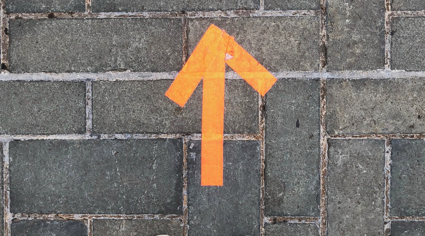 Orange tape arrow pointing up on a brick walkway, indicating direction.