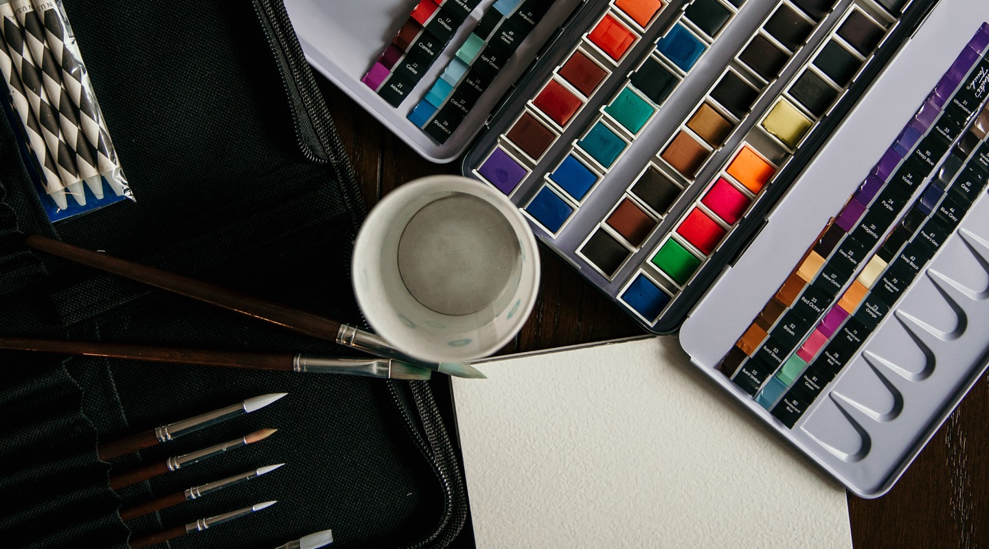 Watercolor paints, brushes, and paper ready for painting, with a cup of water.