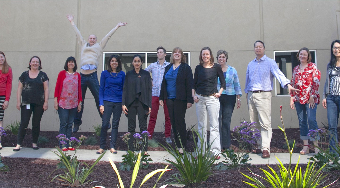 Group photo outside a building with people smiling and jumping. Purple flowers in front.