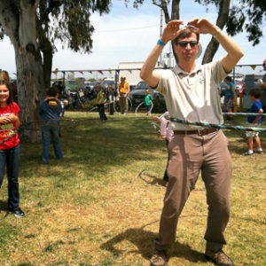 Man trying to hula hoop at a family fun day event. Kids and adults in the background.