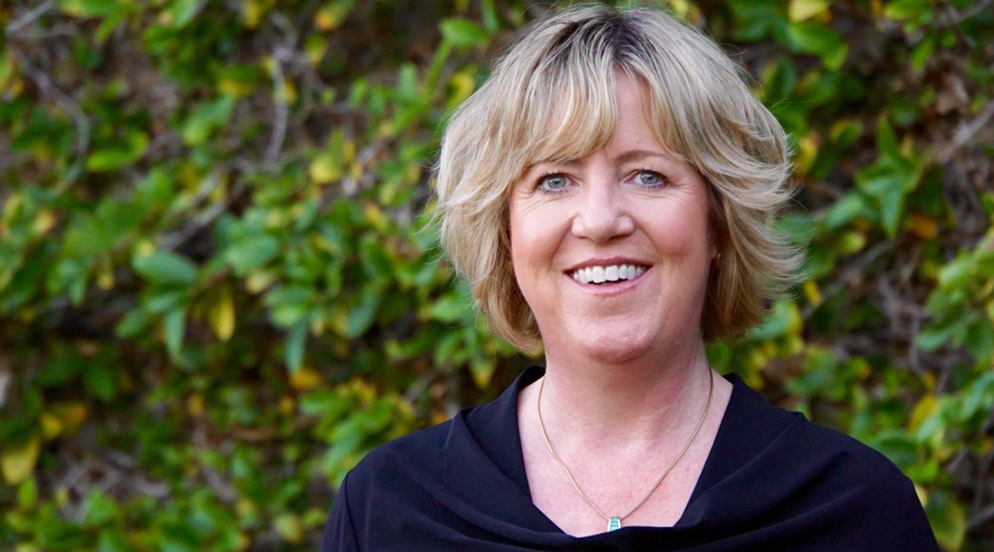 Karen Howard, smiling, with short blonde hair, in front of a green leafy background.