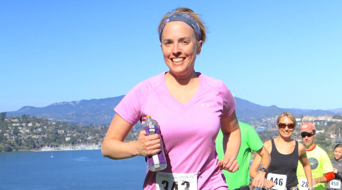 Smiling runner in a race, holding water bottle, with scenic water view in background.
