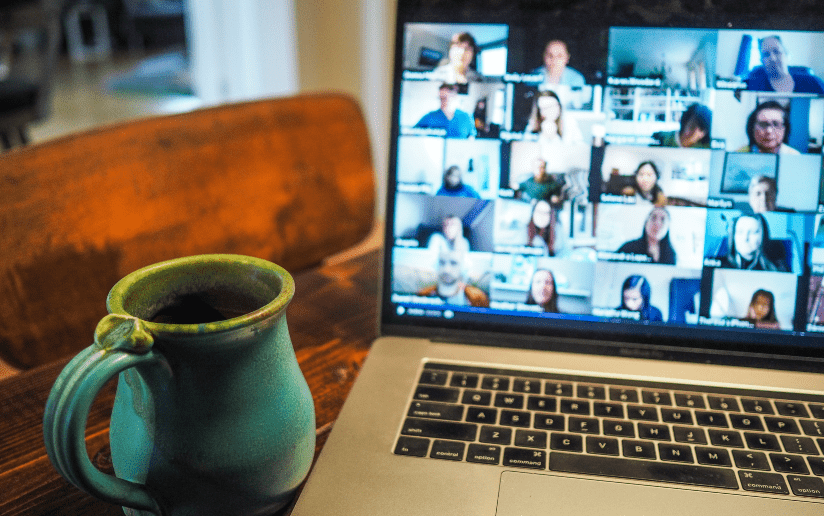 Laptop screen showing a large video conference call with many participants. Coffee mug in foreground.