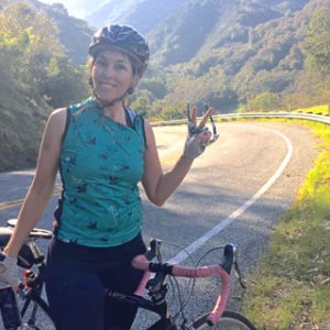 Woman on road bike smiles, giving peace sign, on a winding mountain road. Cycling adventure.