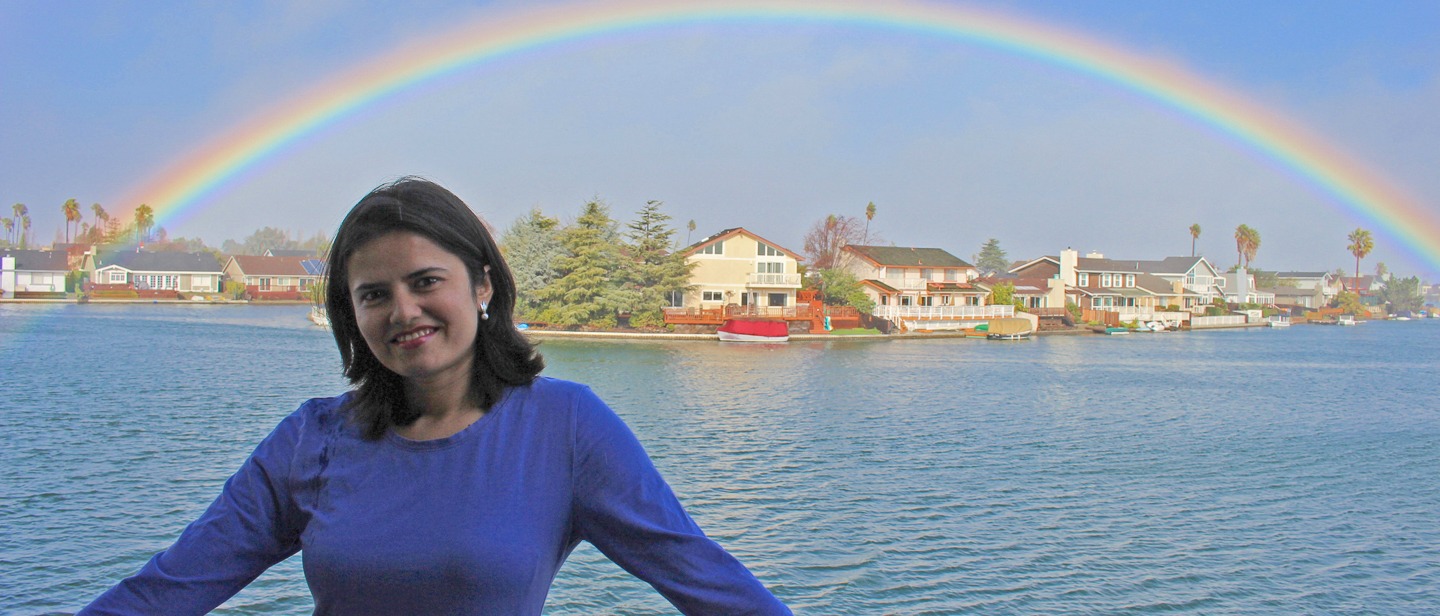 Woman smiles in front of lake with houses and a rainbow arching above.