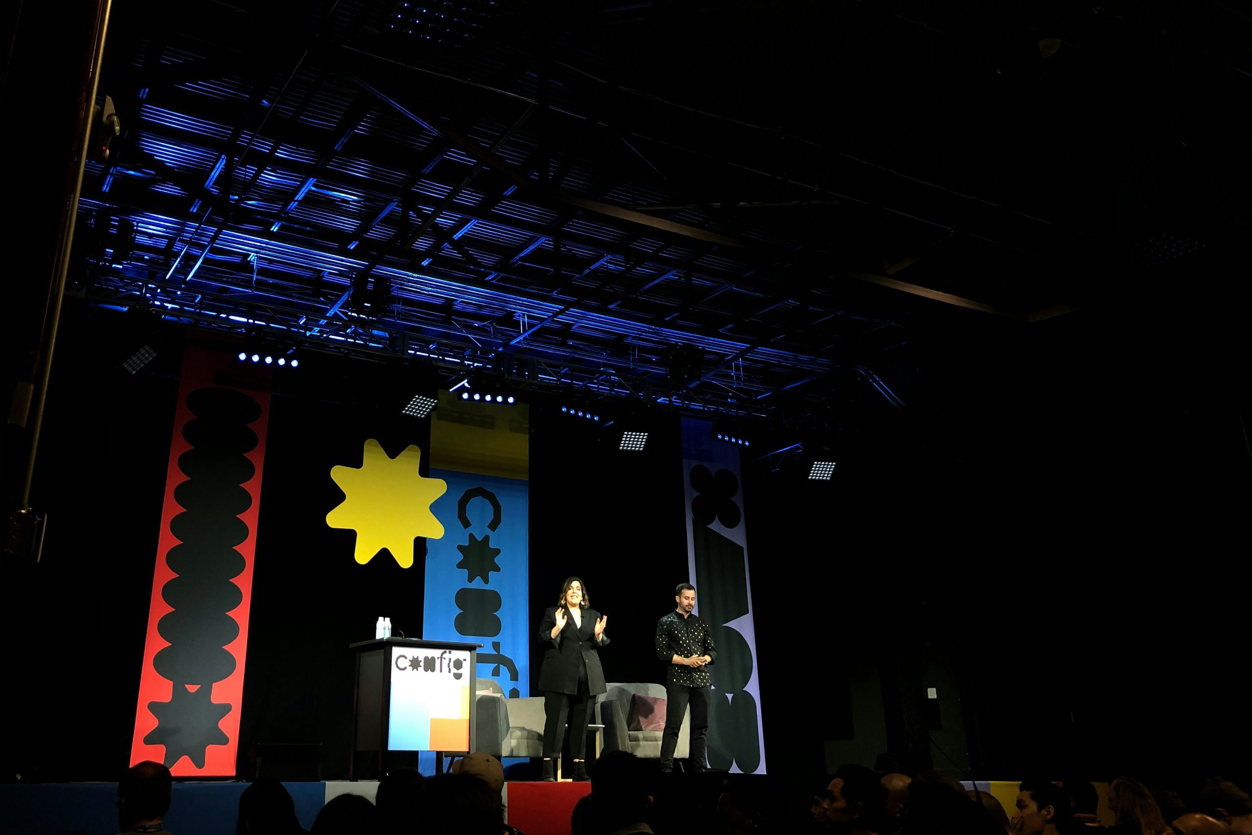 Two speakers on stage at an event with colorful decorations and logo.